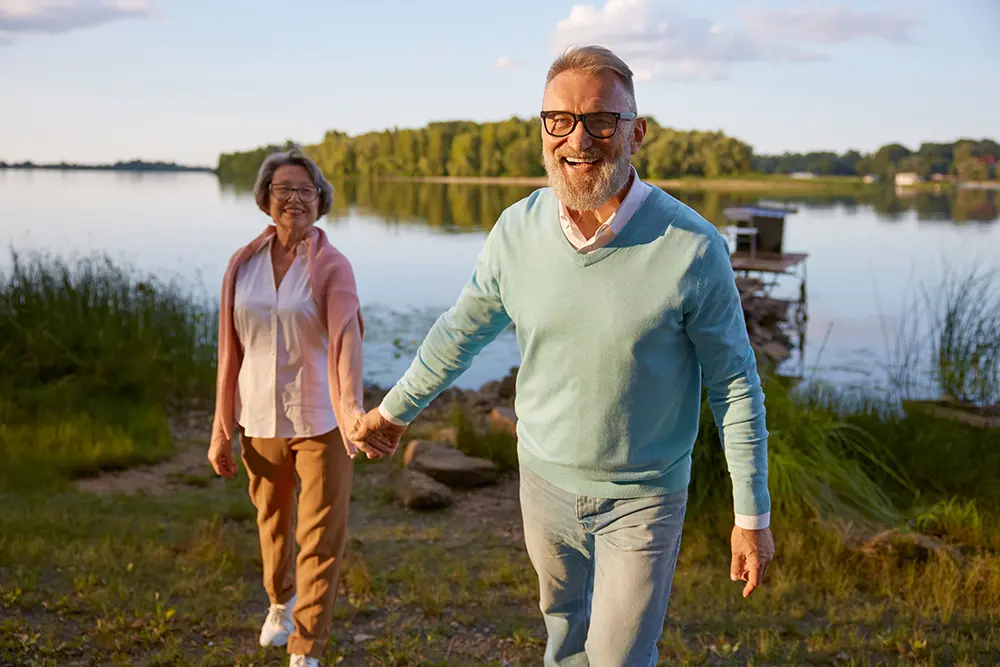 middle age couple walking by river smiling, holding hands